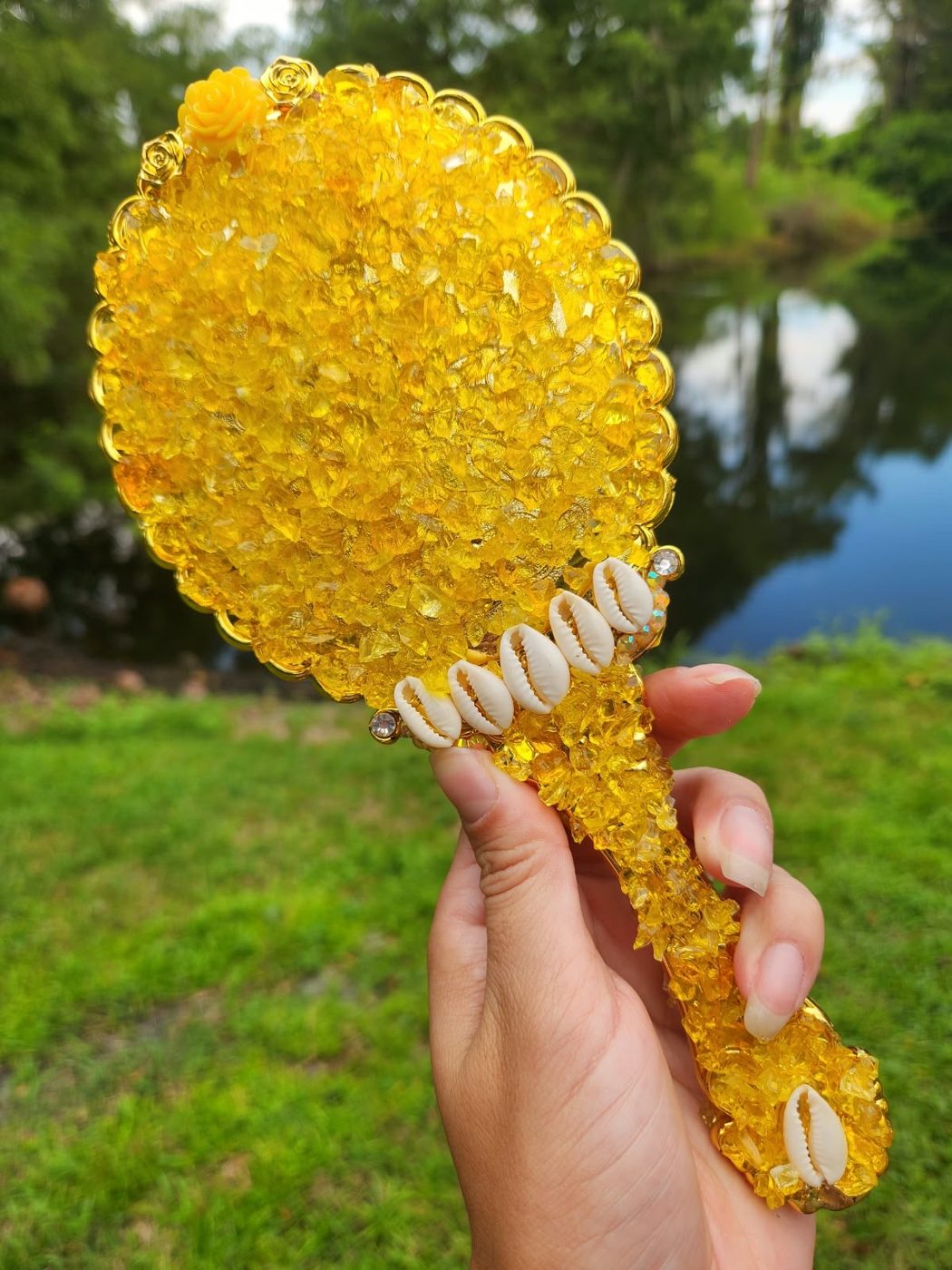 Mão segurando o verso de um espelho artesanal coberto de crushed glass dourado e búzios