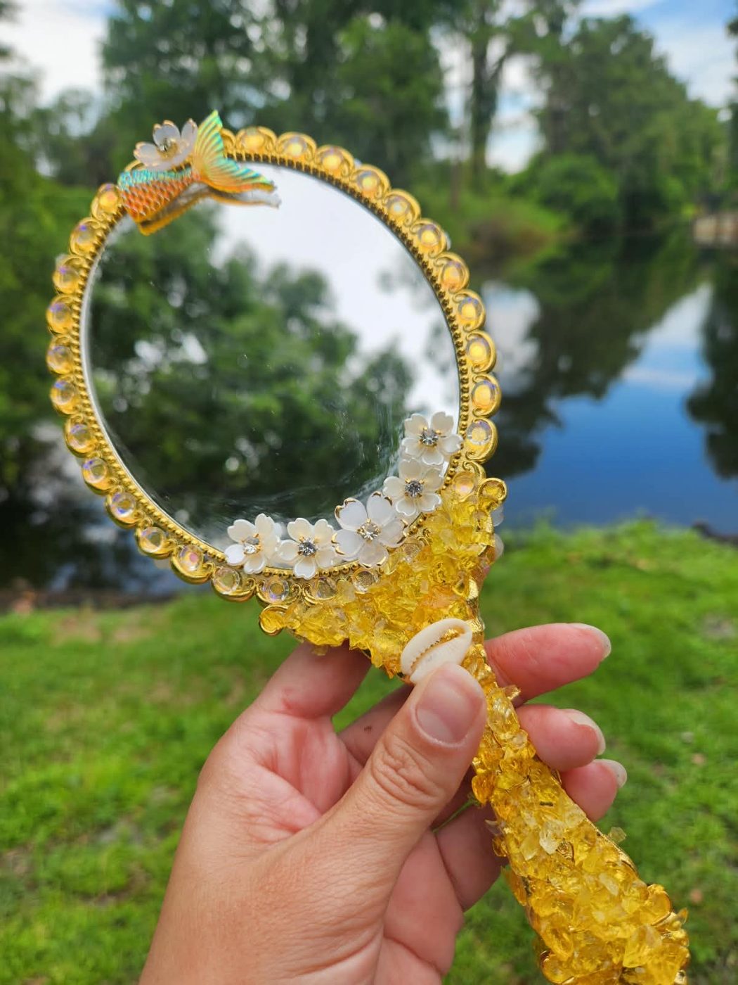 Espelho Reflexo de Areia com flores brancas e miniatura de sereia, diante de um lago