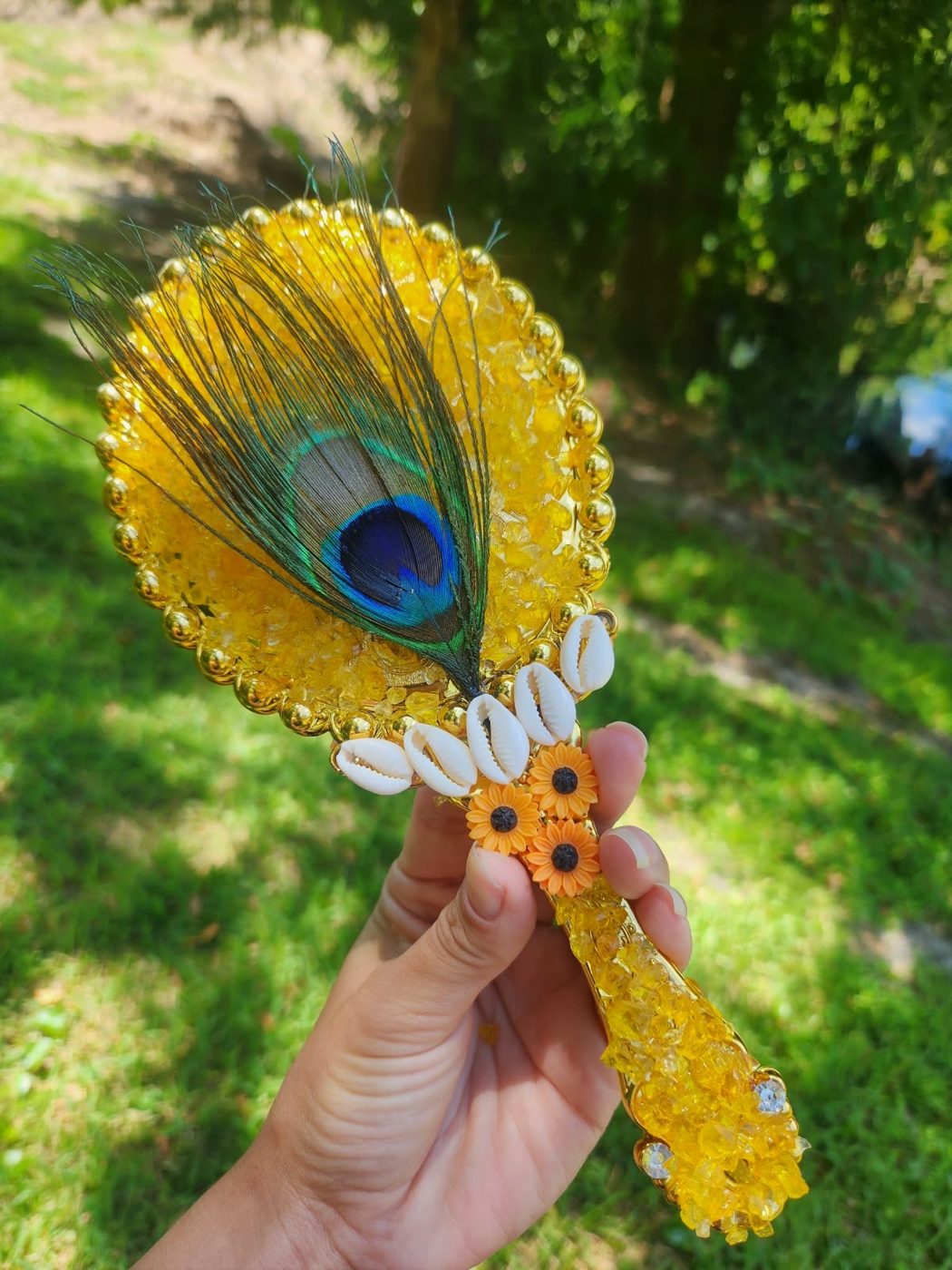 Espelho Reflexo de Areia com pena de pavão, crushed glass dourado e girassóis, nas mãos de uma cliente